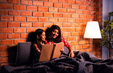 Hintli kızlar gece rahat yorganın altında uyumadan önce masal okurlar. Asyalı küçük kardeş kardeşler birlikte yatakta uzanıp, sıcak yatak odası ortamında gece rutininin tadını çıkarıyorlar.