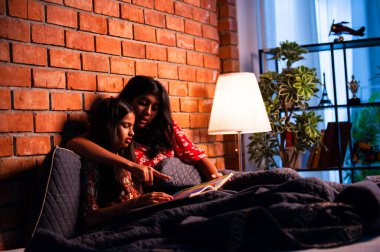 Hintli kızlar gece rahat yorganın altında uyumadan önce masal okurlar. Asyalı küçük kardeş kardeşler birlikte yatakta uzanıp, sıcak yatak odası ortamında gece rutininin tadını çıkarıyorlar.