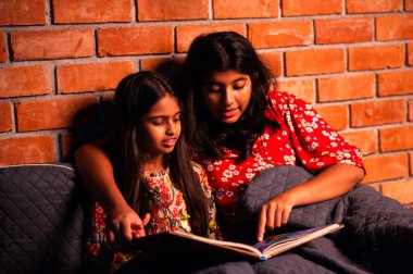 Hintli kızlar gece rahat yorganın altında uyumadan önce masal okurlar. Asyalı küçük kardeş kardeşler birlikte yatakta uzanıp, sıcak yatak odası ortamında gece rutininin tadını çıkarıyorlar.