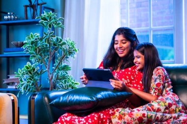 Hintli küçük kız kardeşler tablet bilgisayarda komik videolar izlerken evde oturuyorlar, Asyalı kız kardeşler dijital eğlencenin, çocukluk eğlencesinin, mutluluğun, aile bağlarının tadını çıkarıyorlar.