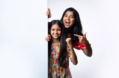 Kızılderili küçük kardeş kızlar, beyaz arkaplana karşı duran, neşeli ifadeli boş tahtayı tanıtıyorlar. Renkli elbiseler giyen Asyalı küçük kız kardeşler reklam mesajı için boş tabela tutuyorlar.