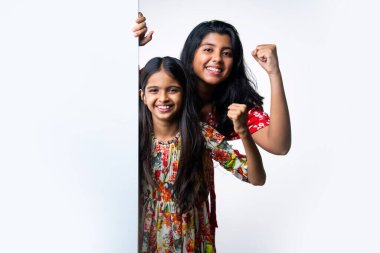 Kızılderili küçük kardeş kızlar, beyaz arkaplana karşı duran, neşeli ifadeli boş tahtayı tanıtıyorlar. Renkli elbiseler giyen Asyalı küçük kız kardeşler reklam mesajı için boş tabela tutuyorlar.