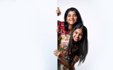 Kızılderili küçük kardeş kızlar, beyaz arkaplana karşı duran, neşeli ifadeli boş tahtayı tanıtıyorlar. Renkli elbiseler giyen Asyalı küçük kız kardeşler reklam mesajı için boş tabela tutuyorlar.