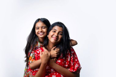 Beyaz arka planda dururken kameraya sarılıp gülümseyen Hintli küçük kardeş kızlar. Renkli elbiseler içinde Asyalı küçük kız kardeşler neşeli ifadelerle sevgi ve neşe gösteriyorlar.