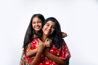 Beyaz arka planda dururken kameraya sarılıp gülümseyen Hintli küçük kardeş kızlar. Renkli elbiseler içinde Asyalı küçük kız kardeşler neşeli ifadelerle sevgi ve neşe gösteriyorlar.