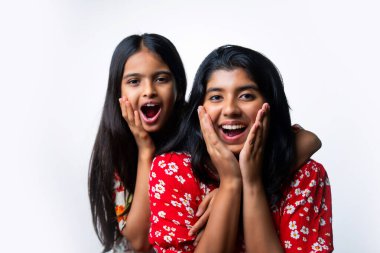 Hintli küçük kız kardeşler, elleri yanaklarında, beyaz arka plana karşı şaşkın ve şaşkın yüz ifadeleri sergiliyorlar, Asyalı genç kardeşler, izole stüdyo ortamında heyecanla tepki veriyorlar.