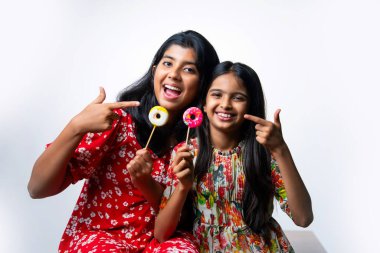 Hintli küçük kardeş lolipop yiyor ve beyaz arka planda otururken şekerlerin tadını çıkarıyor. Renkli elbiseli Asyalı küçük kız kardeşler ellerinde şekerlerle neşeli ifadeler sergiliyorlar.
