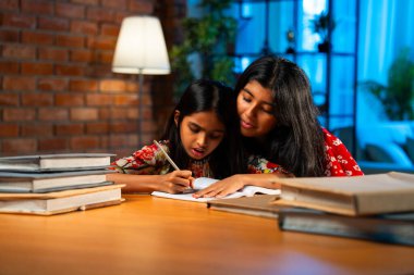 Hintli abla çalışma masasında evde otururken kalemle küçük kız kardeşine ders veriyor. Asyalı küçük kızlar ev ortamında ev ödevi yaparken birlikte öğrenme ve çalışma üzerine odaklandılar.