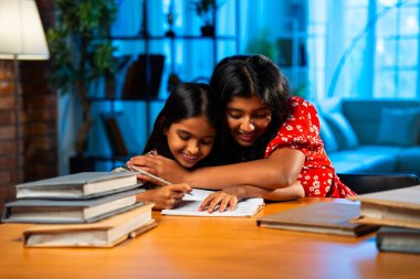 Hintli abla çalışma masasında evde otururken kalemle küçük kız kardeşine ders veriyor. Asyalı küçük kızlar ev ortamında ev ödevi yaparken birlikte öğrenme ve çalışma üzerine odaklandılar.