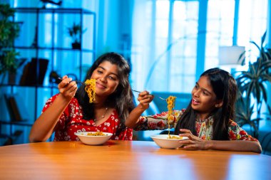 Evde erişte yiyen Hintli küçük kardeş kızlar oturma odasındaki kanepede oturuyorlar. Asyalı küçük kız kardeşler yemek çubuklarıyla, günlük yaşam tarzıyla, mutlulukla ve bağlanmayla vakit geçiriyorlar.