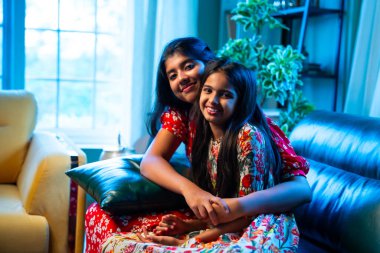 Hintli küçük kız kardeşler kameraya gülümserken kanepede yan yana otururlar, neşeli Asyalı kız kardeşler doğrudan bakar, evde kaynaşırlar, neşeli ifadeler, rahat bir çocukluk anı, mutluluk