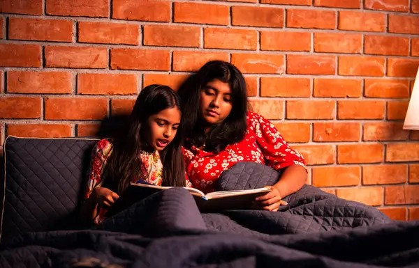 Hintli kızlar gece rahat yorganın altında uyumadan önce masal okurlar. Asyalı küçük kardeş kardeşler birlikte yatakta uzanıp, sıcak yatak odası ortamında gece rutininin tadını çıkarıyorlar.