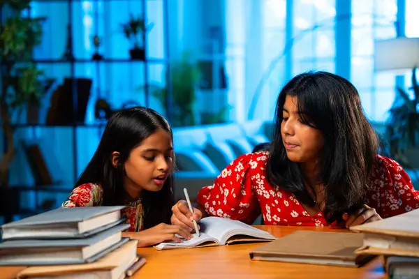 Hintli abla çalışma masasında evde otururken kalemle küçük kız kardeşine ders veriyor. Asyalı küçük kızlar ev ortamında ev ödevi yaparken birlikte öğrenme ve çalışma üzerine odaklandılar.