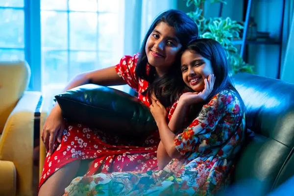 Hintli küçük kız kardeşler kameraya gülümserken kanepede yan yana otururlar, neşeli Asyalı kız kardeşler doğrudan bakar, evde kaynaşırlar, neşeli ifadeler, rahat bir çocukluk anı, mutluluk