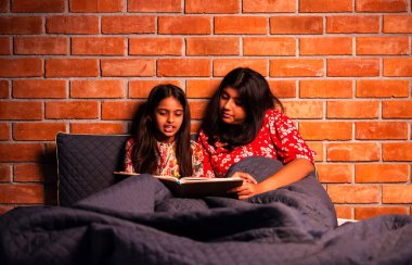 Hintli kızlar gece rahat yorganın altında uyumadan önce masal okurlar. Asyalı küçük kardeş kardeşler birlikte yatakta uzanıp, sıcak yatak odası ortamında gece rutininin tadını çıkarıyorlar.