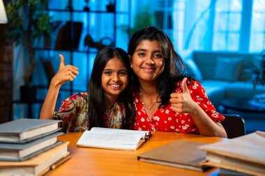 Hintli abla çalışma masasında evde otururken kalemle küçük kız kardeşine ders veriyor. Asyalı küçük kızlar ev ortamında ev ödevi yaparken birlikte öğrenme ve çalışma üzerine odaklandılar.