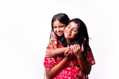 Beyaz arka planda dururken kameraya sarılıp gülümseyen Hintli küçük kardeş kızlar. Renkli elbiseler içinde Asyalı küçük kız kardeşler neşeli ifadelerle sevgi ve neşe gösteriyorlar.