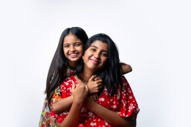 Beyaz arka planda dururken kameraya sarılıp gülümseyen Hintli küçük kardeş kızlar. Renkli elbiseler içinde Asyalı küçük kız kardeşler neşeli ifadelerle sevgi ve neşe gösteriyorlar.