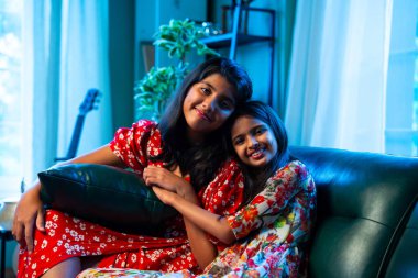 Hintli küçük kız kardeşler kameraya gülümserken kanepede yan yana otururlar, neşeli Asyalı kız kardeşler doğrudan bakar, evde kaynaşırlar, neşeli ifadeler, rahat bir çocukluk anı, mutluluk