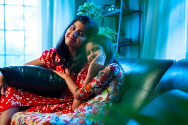 Hintli küçük kız kardeşler kameraya gülümserken kanepede yan yana otururlar, neşeli Asyalı kız kardeşler doğrudan bakar, evde kaynaşırlar, neşeli ifadeler, rahat bir çocukluk anı, mutluluk