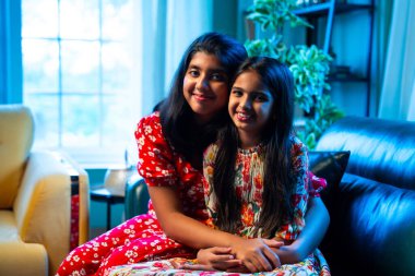 Hintli küçük kız kardeşler kameraya gülümserken kanepede yan yana otururlar, neşeli Asyalı kız kardeşler doğrudan bakar, evde kaynaşırlar, neşeli ifadeler, rahat bir çocukluk anı, mutluluk