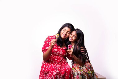Hintli küçük kardeş lolipop yiyor ve beyaz arka planda otururken şekerlerin tadını çıkarıyor. Renkli elbiseli Asyalı küçük kız kardeşler ellerinde şekerlerle neşeli ifadeler sergiliyorlar.