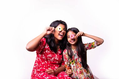 Hintli küçük kardeş lolipop yiyor ve beyaz arka planda otururken şekerlerin tadını çıkarıyor. Renkli elbiseli Asyalı küçük kız kardeşler ellerinde şekerlerle neşeli ifadeler sergiliyorlar.