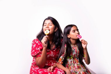 Hintli küçük kardeş lolipop yiyor ve beyaz arka planda otururken şekerlerin tadını çıkarıyor. Renkli elbiseli Asyalı küçük kız kardeşler ellerinde şekerlerle neşeli ifadeler sergiliyorlar.