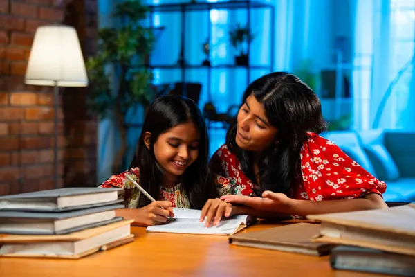Hintli abla çalışma masasında evde otururken kalemle küçük kız kardeşine ders veriyor. Asyalı küçük kızlar ev ortamında ev ödevi yaparken birlikte öğrenme ve çalışma üzerine odaklandılar.
