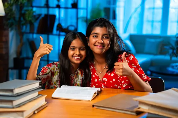 Hintli abla çalışma masasında evde otururken kalemle küçük kız kardeşine ders veriyor. Asyalı küçük kızlar ev ortamında ev ödevi yaparken birlikte öğrenme ve çalışma üzerine odaklandılar.