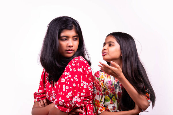 Индийские сестрички дерутся и сидят спиной к спине со злобными выражениями на белом фоне. Маленькие азиатки в красочных платьях показывают конфликты, перепады настроения и эмоции
