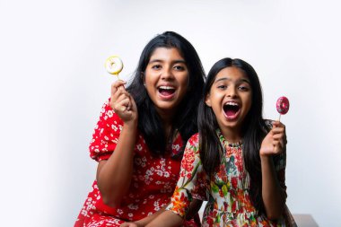 Hintli küçük kardeş lolipop yiyor ve beyaz arka planda otururken şekerlerin tadını çıkarıyor. Renkli elbiseli Asyalı küçük kız kardeşler ellerinde şekerlerle neşeli ifadeler sergiliyorlar.