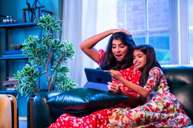 Hintli küçük kız kardeşler tablet bilgisayarda komik videolar izlerken evde oturuyorlar, Asyalı kız kardeşler dijital eğlencenin, çocukluk eğlencesinin, mutluluğun, aile bağlarının tadını çıkarıyorlar.