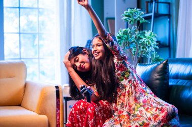 Hintli küçük kız kardeşler evdeki kanepede otururken video oyunu oynuyorlar, Asyalı kardeş kızlar joystickle oyun oynuyorlar, neşeli çocukluk eğlenceleri, elektronik eğlence, aile bağları.