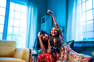 Hintli küçük kız kardeşler evdeki kanepede otururken video oyunu oynuyorlar, Asyalı kardeş kızlar joystickle oyun oynuyorlar, neşeli çocukluk eğlenceleri, elektronik eğlence, aile bağları.