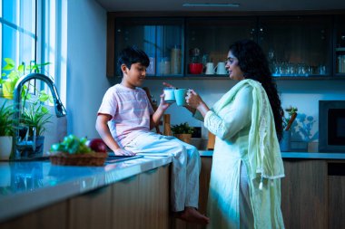 Hintli bir çocuk modern mutfakta genç bir anneyle birlikte camdan süt içiyor, evde birlikte kaliteli zaman geçiriyorlar, günlük rutin, mutlu ebeveyn çocuk bağı videosu.