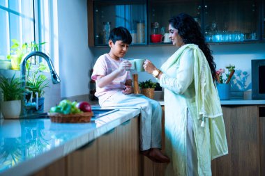 Hintli bir çocuk modern mutfakta genç bir anneyle birlikte camdan süt içiyor, evde birlikte kaliteli zaman geçiriyorlar, günlük rutin, mutlu ebeveyn çocuk bağı videosu.