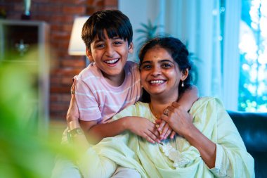 Hintli anne küçük çocuk kanepenin üzerinde modern evde sarılıyor, birlikte iyi vakit geçiriyorlar, sevgi ve sıcaklık gösteriyorlar, ebeveyn-çocuk ilişkisi yaşam tarzı aile mutluluğu kavramı.