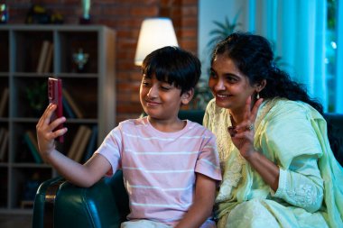 Hintli çocuk, annesiyle selfie çekiyor. Modern evde, kanepenin üzerinde akıllı telefon kullanıyor. Birlikte zaman geçiriyorlar. Şefkat, sevgi, neşe, sıcaklık, aile içi çocuk ilişkileri.