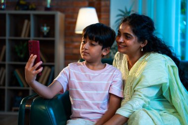 Hintli çocuk, annesiyle selfie çekiyor. Modern evde, kanepenin üzerinde akıllı telefon kullanıyor. Birlikte zaman geçiriyorlar. Şefkat, sevgi, neşe, sıcaklık, aile içi çocuk ilişkileri.