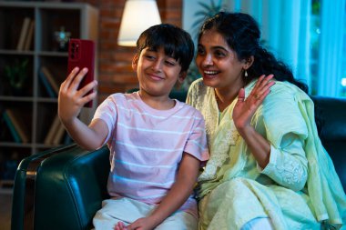 Hintli çocuk ve anne akıllı telefondan video çağrısında bulundular. Modern bir evde, koltukta oturuyorlar, birlikte kaliteli zaman geçiriyorlar. Şefkat, sevgi, aile içi çocuk ilişkisi gösteriyorlar.