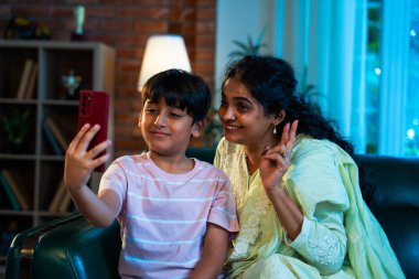 Hintli çocuk, annesiyle selfie çekiyor. Modern evde, kanepenin üzerinde akıllı telefon kullanıyor. Birlikte zaman geçiriyorlar. Şefkat, sevgi, neşe, sıcaklık, aile içi çocuk ilişkileri.