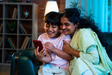 Hintli çocuk ve anne akıllı telefondan sörf yaparken modern evde kanepede oturuyorlar, birlikte kaliteli zaman geçiriyorlar, teknolojiyi kullanıyorlar, neşeyi paylaşıyorlar, ebeveynlik ilişkileri, aile ilişkileri.