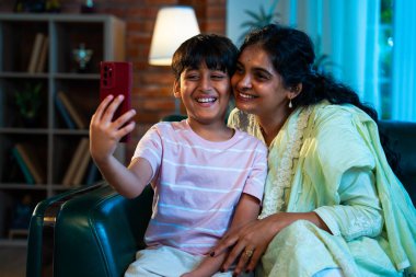 Hintli çocuk ve anne akıllı telefondan video çağrısında bulundular. Modern bir evde, koltukta oturuyorlar, birlikte kaliteli zaman geçiriyorlar. Şefkat, sevgi, aile içi çocuk ilişkisi gösteriyorlar.
