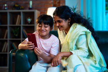 Hintli çocuk ve anne akıllı telefondan sörf yaparken modern evde kanepede oturuyorlar, birlikte kaliteli zaman geçiriyorlar, teknolojiyi kullanıyorlar, neşeyi paylaşıyorlar, ebeveynlik ilişkileri, aile ilişkileri.