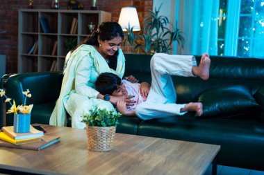 Hintli çocuk ve anne gülerek modern evde kanepede oturuyorlar, birlikte kaliteli zaman geçiriyorlar, şefkat gösteriyorlar, sevgi, neşe, mutluluk, ebeveyn-çocuk ilişkisi yaşam tarzı kapalı