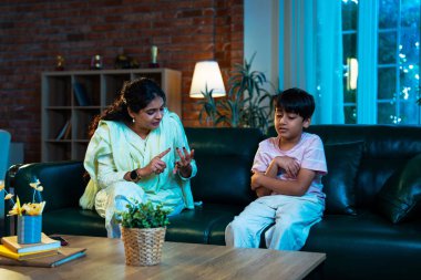 Hintli anne oğlunu azarlıyor, modern evde koltukta otururken, iyi alışkanlıklar öğretiyor, disiplini teşvik ediyor, kaliteli zaman geçirtiyor, çocuğa sevgiyle yol gösteriyor, ebeveyn çocuk ilişkisini kapalı alanda gösteriyor.