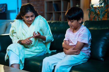 Hintli anne oğlunu azarlıyor, modern evde koltukta otururken, iyi alışkanlıklar öğretiyor, disiplini teşvik ediyor, kaliteli zaman geçirtiyor, çocuğa sevgiyle yol gösteriyor, ebeveyn çocuk ilişkisini kapalı alanda gösteriyor.