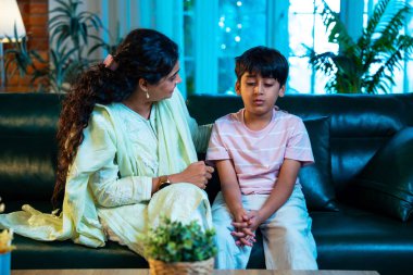 Hintli anne oğlunu azarlıyor, modern evde koltukta otururken, iyi alışkanlıklar öğretiyor, disiplini teşvik ediyor, kaliteli zaman geçirtiyor, çocuğa sevgiyle yol gösteriyor, ebeveyn çocuk ilişkisini kapalı alanda gösteriyor.