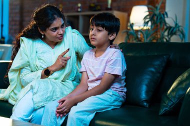 Hintli anne oğlunu azarlıyor, modern evde koltukta otururken, iyi alışkanlıklar öğretiyor, disiplini teşvik ediyor, kaliteli zaman geçirtiyor, çocuğa sevgiyle yol gösteriyor, ebeveyn çocuk ilişkisini kapalı alanda gösteriyor.